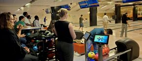 A group of people enjoying bowling. Several screens display scores. Bowling balls are arranged on racks.