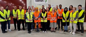 A group of people wearing yellow and orange high-visibility vests stand outside an office building with Valmet signage.