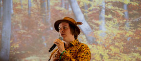 A person wearing a colourful shirt and brown hat speaks into a microphone with a background of an image of a forest.