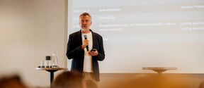 A man in a black blazer speaks using a microphone in front of a projector screen during a presentation.