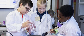Three children wearing lab coats conducting a science experiment with purple substance on a tray.