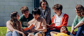 Group of friends sitting on grass reading books, near a stone building.