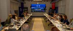 A conference room with attendees seated around a U-shaped table, facing a screen displaying 'SUN' and 'WELCOME'.