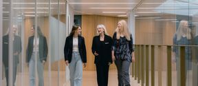 Three women walking through a modern corridor with glass walls, dressed in casual and business attire.