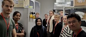Group of people standing in a room with shelves and boxes, many wearing red lanyards and black or striped tops.