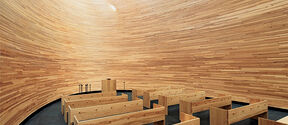 Modern chapel with curved wooden walls, wooden benches, and an altar under a round, bright ceiling.