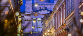 Person in winter clothes walking on a festive street with Christmas lights, leading to the white Helsinki Cathedral in evening.