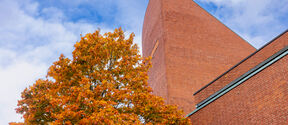 A brick building with a tall pointed roof beside an autumnal tree under a partly cloudy sky.