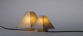 Two table lamps with algae-paper shades and wooden bases. Both are lit and connected by a black cord.