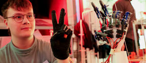 A person wearing a grey shirt and black glove shows a victory sign next to a robotic hand in a bright room.