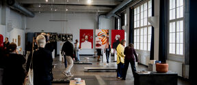 People in an art gallery with large windows, viewing various artworks including portraits and abstract pieces.