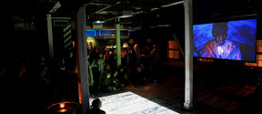 A dimly-lit nightclub event space, with projections of a ﬁlm on a screen and water waves on the ﬂoor as the audience watches.