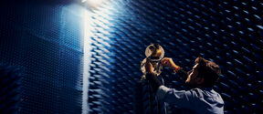 Person adjusts equipment in an anechoic chamber with blue acoustic foam wedges and bright overhead light.