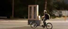 A person rides a cargo bike carrying a large wooden cabinet at night.
