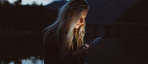 A person with long blonde hair is looking at a phone screen beside a lake during dusk.
