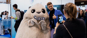 A person poses with a large white furry mascot holding a sign that says 'Sievo' at an event. Another person takes a photo.