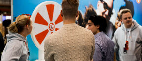 People standing around a red and white spinning wheel at an event. A man in a beige sweater is in the foreground.