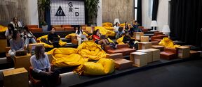 People sitting on tiered seating with yellow bean bags and wooden cubes in a modern room with concrete walls.