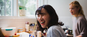 Two people in a kitchen with various items on the counter. One person is holding a straw.