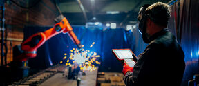 Työntekijä käyttää tablettia, kun robottikäsi hitsaa metallia ja kipinöitä lentää teollisuusympäristössä.