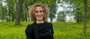 Eeva Furman in a black dress and gloves standing in a field with yellow flowers and trees in the background.