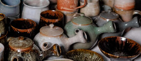 A collection of ceramic mugs, teapots, and bowls in various earthy tones, including green, brown, and beige.