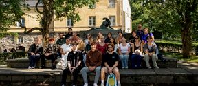 A group of people sitting on stone steps in a park with trees and a yellow building in the background.