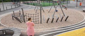 A woman in a striped dress watches children playing on a playground with wooden structures and a net.