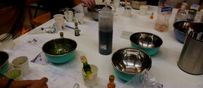A table with bowls, bottles, and cups. People are working with various items, including a bottle of dark liquid.