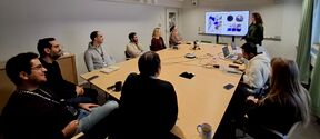 A group of people in a meeting room watching a presentation on a large screen. Laptops and coffee mugs are on the table.