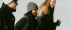 Three people in winter clothing walking outdoors on a snowy day. One holds a coffee cup.