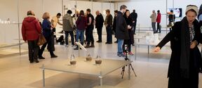 People are gathered in a modern exhibition space with tables displaying various items. Some are interacting with the exhibits.