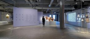 A modern art gallery with pottery, hanging art, and a projection. A person walks past a lavender wall with text.