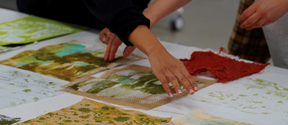 Two people arranging various textured and coloured fabric samples on a white table.