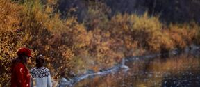 Two people walking by a river in autumn. One wears a red coat and hat, the other a white sweater with patterns.