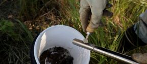A gloved hand holds a tool over a metal pipe, with a white bucket containing soil in the background.