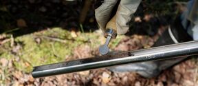 A gloved hand holds a soil sampler with soil inside, over a forest floor covered in leaves and moss.