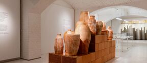 An art gallery with large ceramic vases on a tiled pedestal. White arched ceiling and walls with text panels.