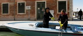 Two people on a boat collecting water samples near a brick building with green shutters.