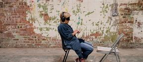 A person wearing headphones sits on a chair facing a folding chair in a room with a worn brick and plaster wall.