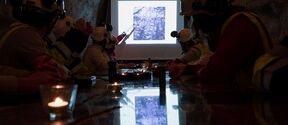 People in helmets and vests sit in a cave-like room, watching a presentation on a screen. A person points at the screen.