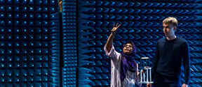 Two people in an anechoic chamber with blue acoustic foam panels. One person gestures while the other holds equipment.
