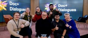A group of people giving thumbs up in front of screens displaying 'Doc+ Dialogues'. Chairs and wooden walls are in the background.