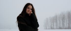 Pitkähiuksinen henkilö tummassa neuleessa seisoo lumisella pellolla, taustalla paljaita puita.