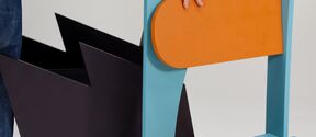 A person touches an abstract chair with orange and blue elements. Black angular shapes are in the background.