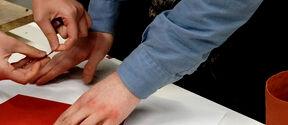 Two people working with red and white paper on a table. One wears a blue shirt, the other has a bracelet.