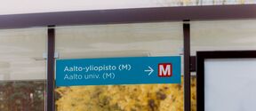 kaksi nuorta istumassa bussipysäkillä, jossa on nuoli Aalto-yliopiston metro-asemalle