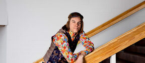 Person leaning on wooden staircase rail, wearing colourful floral shirt, patterned waistcoat, and a headband.