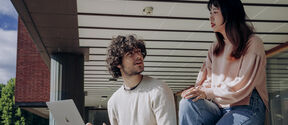 Two students outside a brick building, one using a silver laptop while talking with the other on a red bench