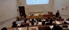 Lecture hall with students watching a man present slides on electrically heated textiles at the front